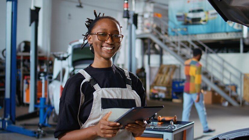 Mechanic holding a tablet in an auto repair shop with various tools and cars in the background.