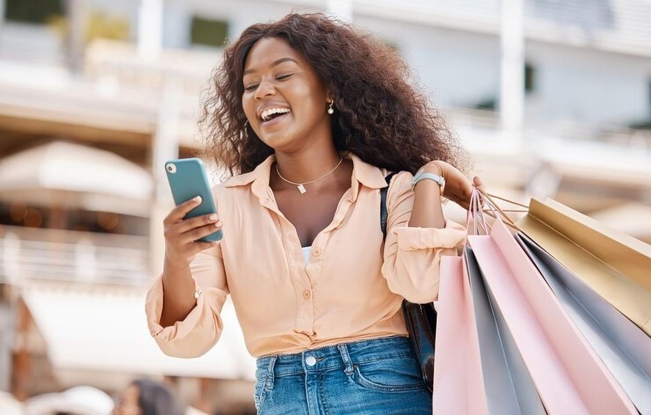 Woman smiling and holding shopping bags while looking at her smartphone outdoors.