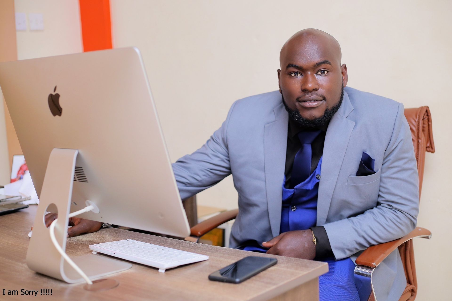 Man in a blue suit seated at a desk with an Apple computer and smartphone.