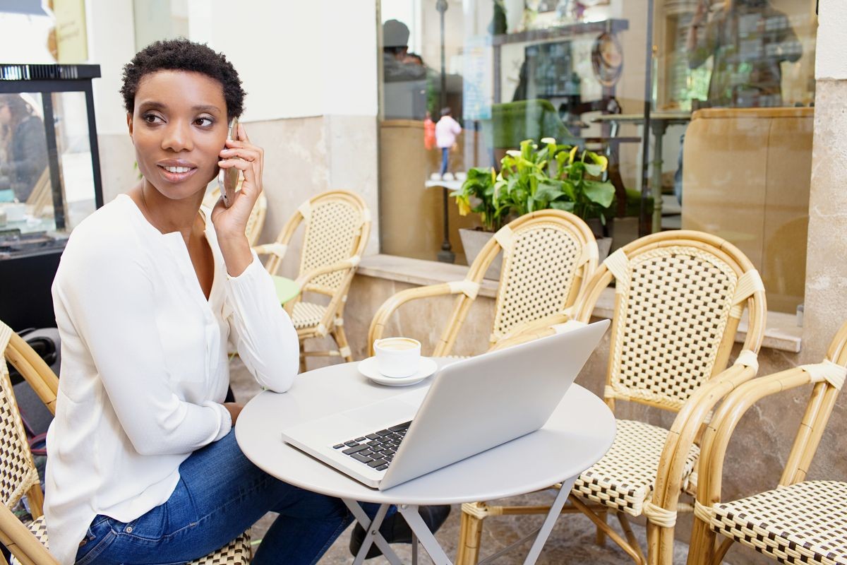 Portrait of professional african american office woman in coffee shop using laptop computer, smart phone call conversation, working in city outdoors. Black business woman using technology, exterior.